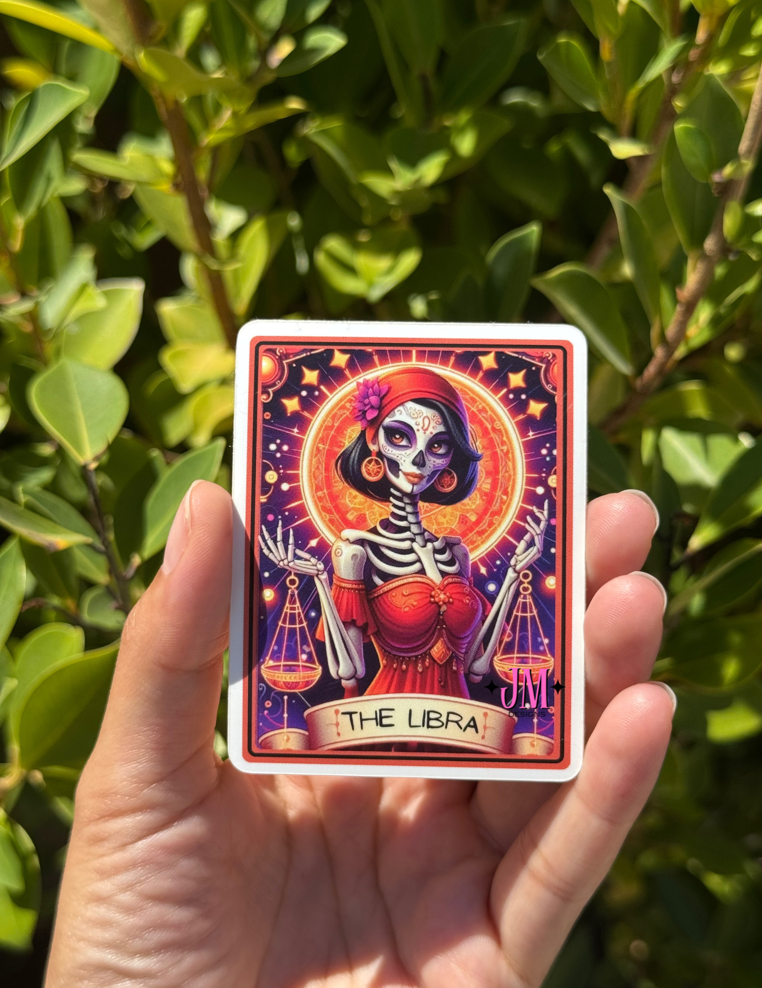 A hand holds a square art card featuring a Day of the Dead skeleton illustration labeled 'THE LIBRA' against a background of green outdoor foliage.